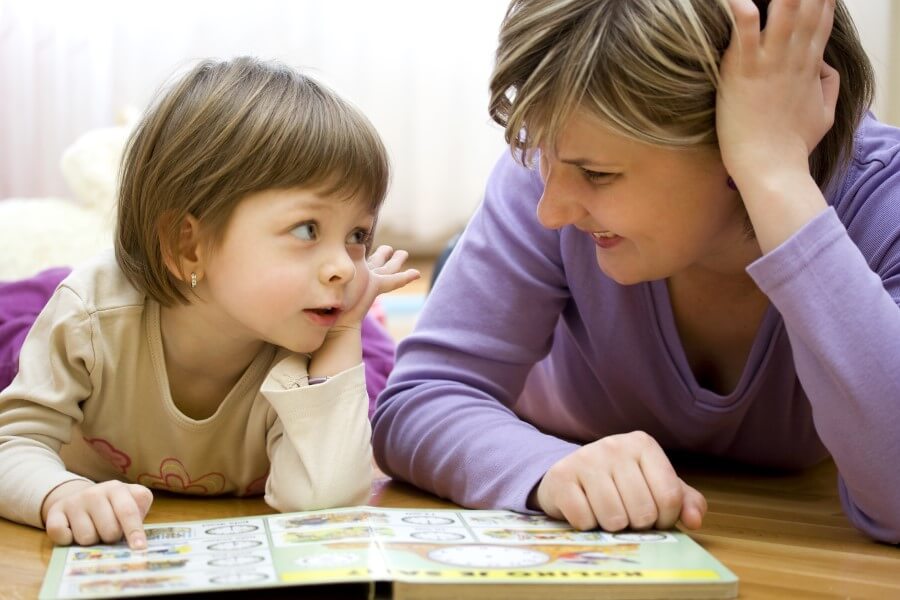 A mature Caucasian mother reads with her young child while both lie on the floor, smiling.