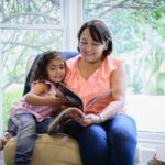 Young Latin American granddaughter sharing a chair with her grandmother and looking at a picture book together.