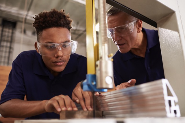 A Caucasian carpenter trains a black high school student on a wood-working machine.
