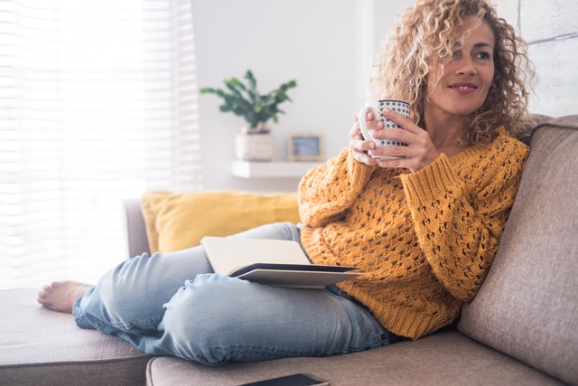 A Caucasian woman sits down on the sofa drinking tea.