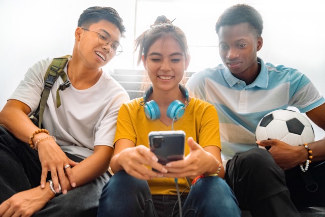 Multiracial group of teen high school classmates sit on stairs looking at teenage girl cell phone.
