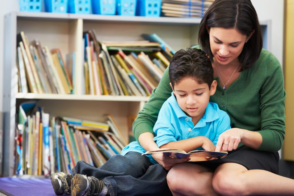 Elementary Pupil Reading With Teacher In Classroom Sitting Down On Floor