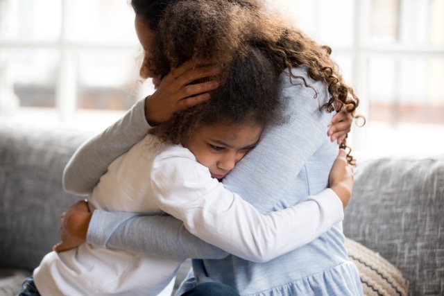 Black mother embraces her frustrated preschooler as they sit on couch together at home.