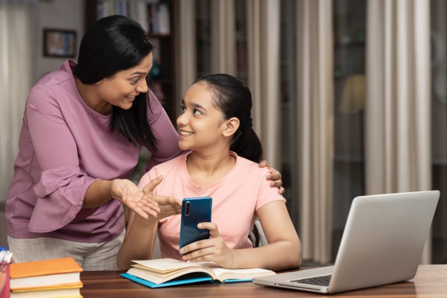 An Southeast Asian teenager shows her mother something on her phone as she does homework. Both are smiling.
