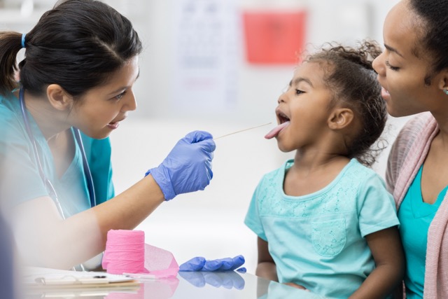 An Asian female pediatrician uses a tongue depressor to look at young African American girl's throat. The girl's mother is holding her.