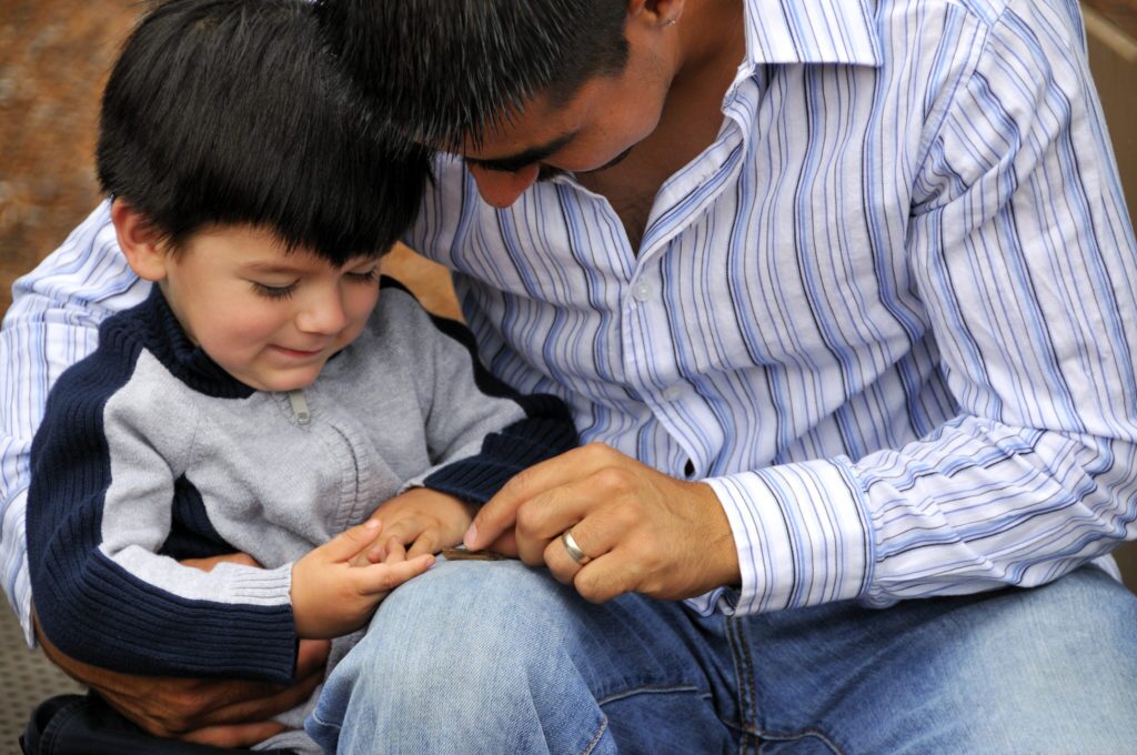 A young father hugs his toddler son as they examine the toddler's fingers with curiosity.