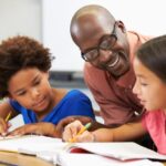 A black teacher smiles as he helps young multiethnic students with their work.