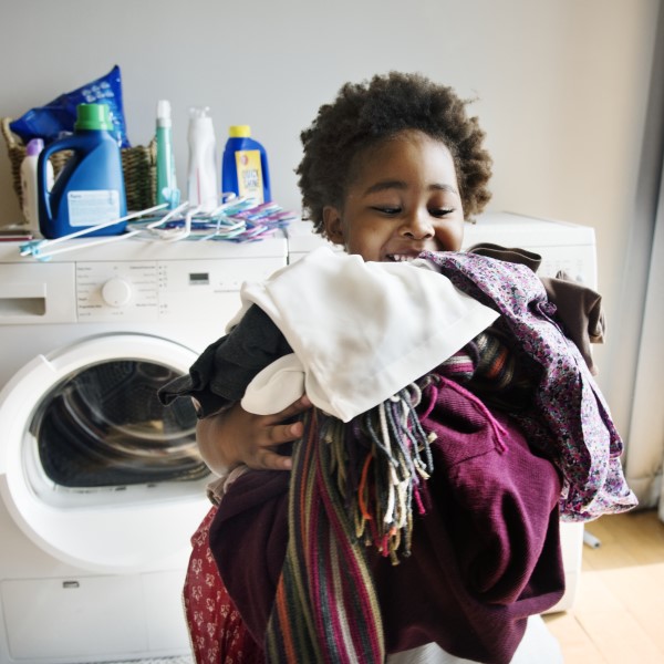 A smiling young black girl carries an armful of laundry from the washing machine.