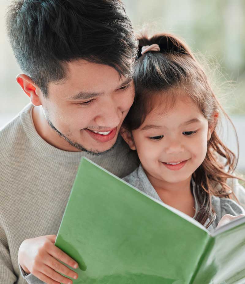 An Asian father reads a book to his toddler daughter as they smile together.