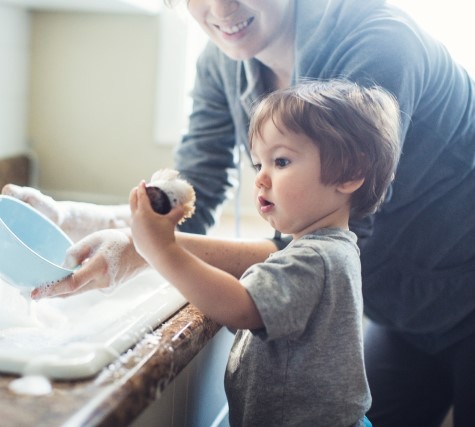 A toddler stands with his mother at the kitchen sink, holding an object with curiosity.