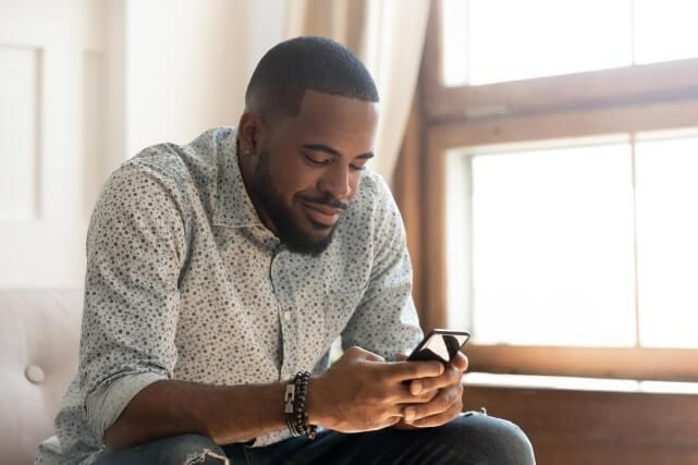 A Black father smiles as he uses his phone while sitting on a couch at home.