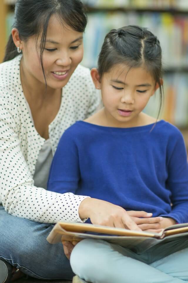 An Asian mother and her elementary school aged daughter read a book together at the library.