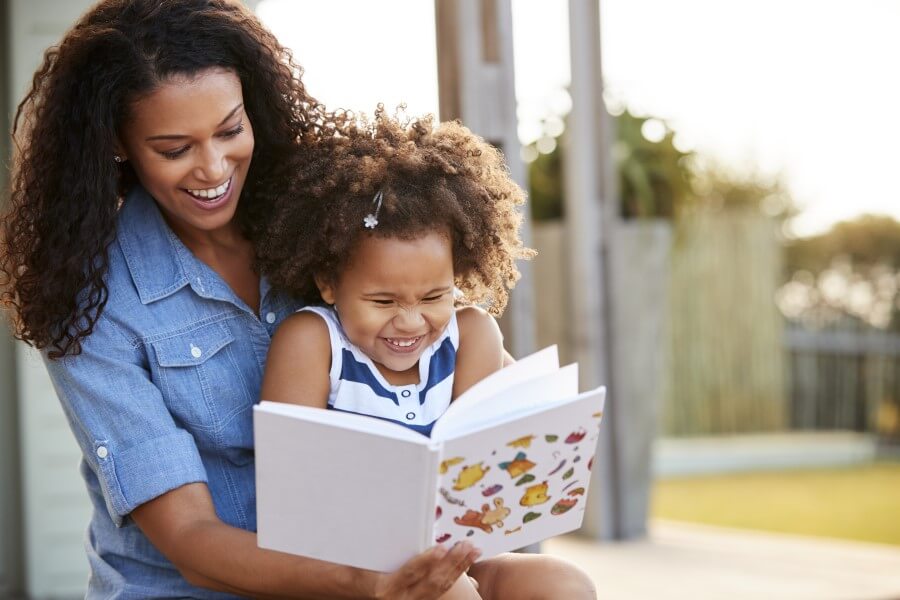 Young black preschooler reading a book while sitting on her mother's lap outside.