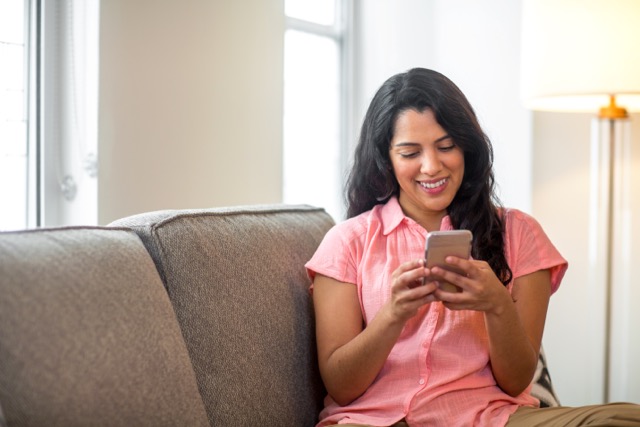 A Latina woman smiles as she reads a text message on her cellphone at home.
