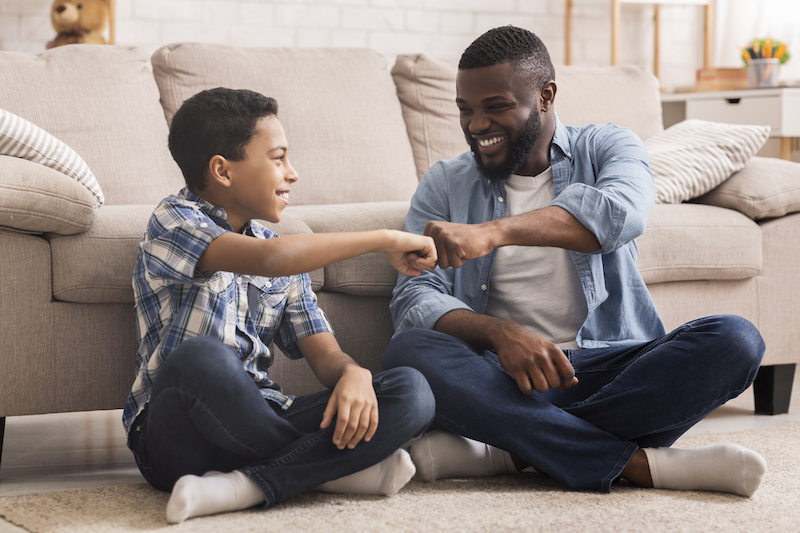 A Black father and his preteen son sit together at home, fist-pumping and smiling. 