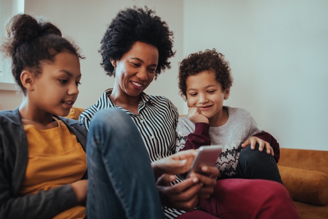 A black mother sits with her young children on a couch, smiling as she reads a ParentPowered message on her cell phone.