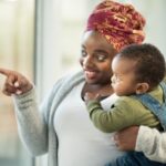 A black mother smiles and points out the window to show her baby something interesting. 