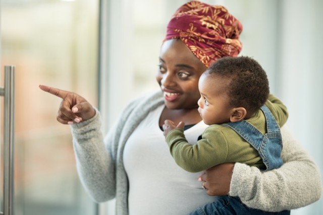 A black mother smiles and points out the window to show her baby something interesting.