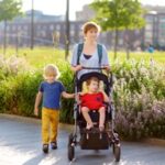 A white woman walks through a park, pushing a stroller with a child that has a disability while his sibling walks near by.