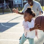 A black grandfather plays basketball with  his young granddaughter in the park.