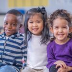 Three multi-ethnic preschool children smile at the camera and put their arms around each other.