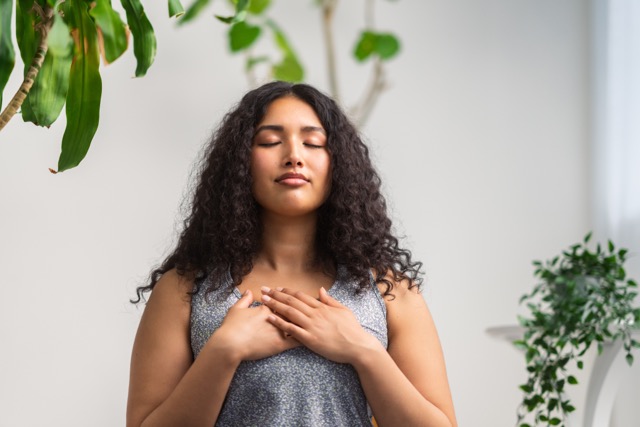 A multiracial woman performs a breathing exercise at home as an act of parent self care.