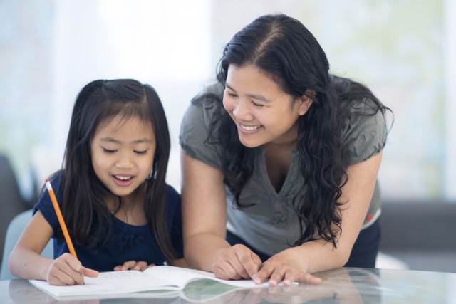 An Asian mother and her young daughter work on homework together while smiling.
