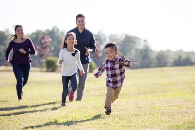 A family of four are chasing each other and playing tag at the park. They are running across the grass and are smiling happily.