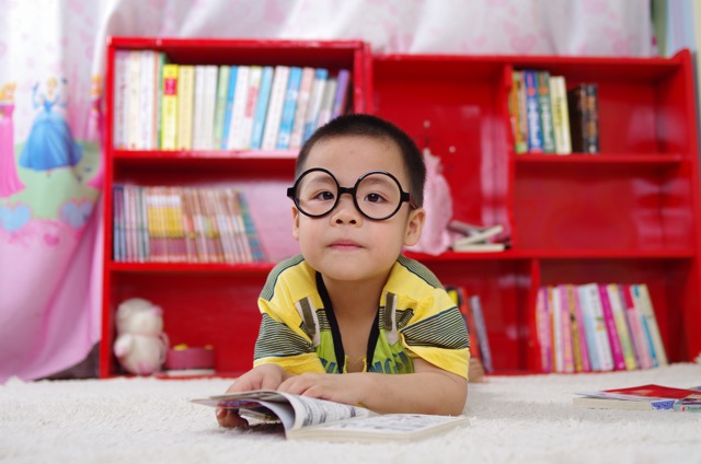 A young Asian boy with glasses looks at the camera as he opens a book at a library.