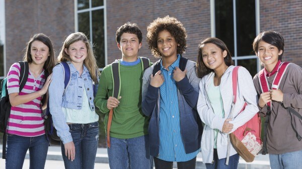 A diverse group of middle school students smile at the camera in front of their school building.