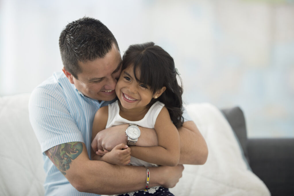 A Hispanic father hugs his preschool daughter as they both smile.
