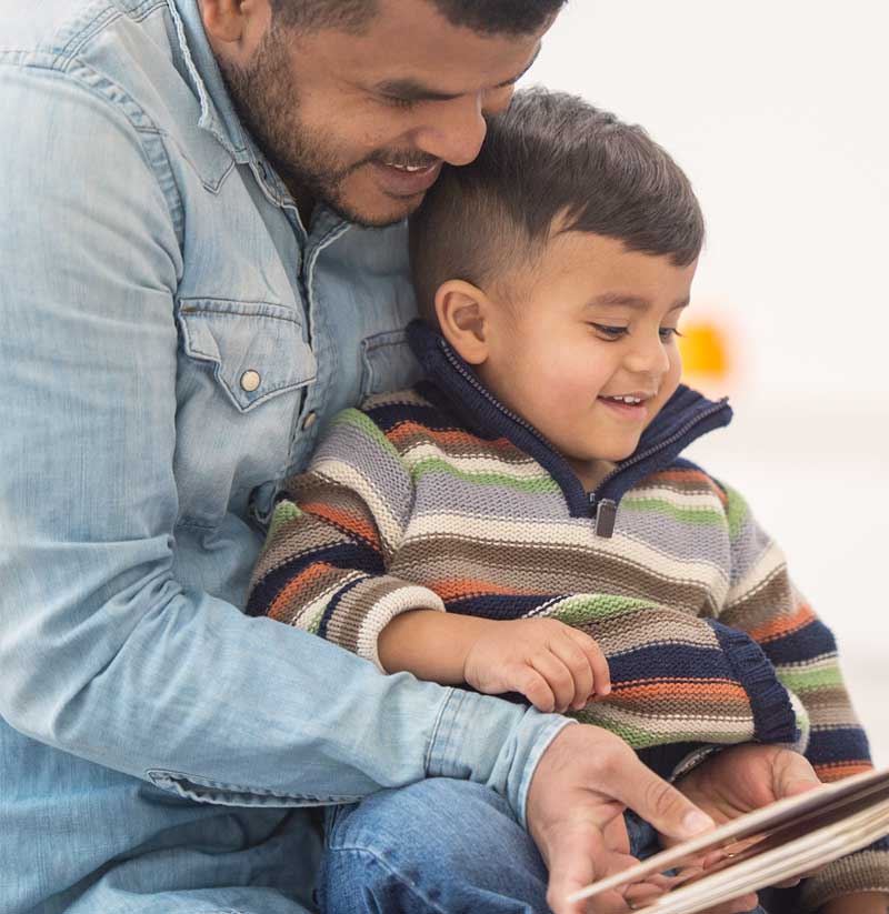 A father reads from a book to his toddler son, while both smile. 