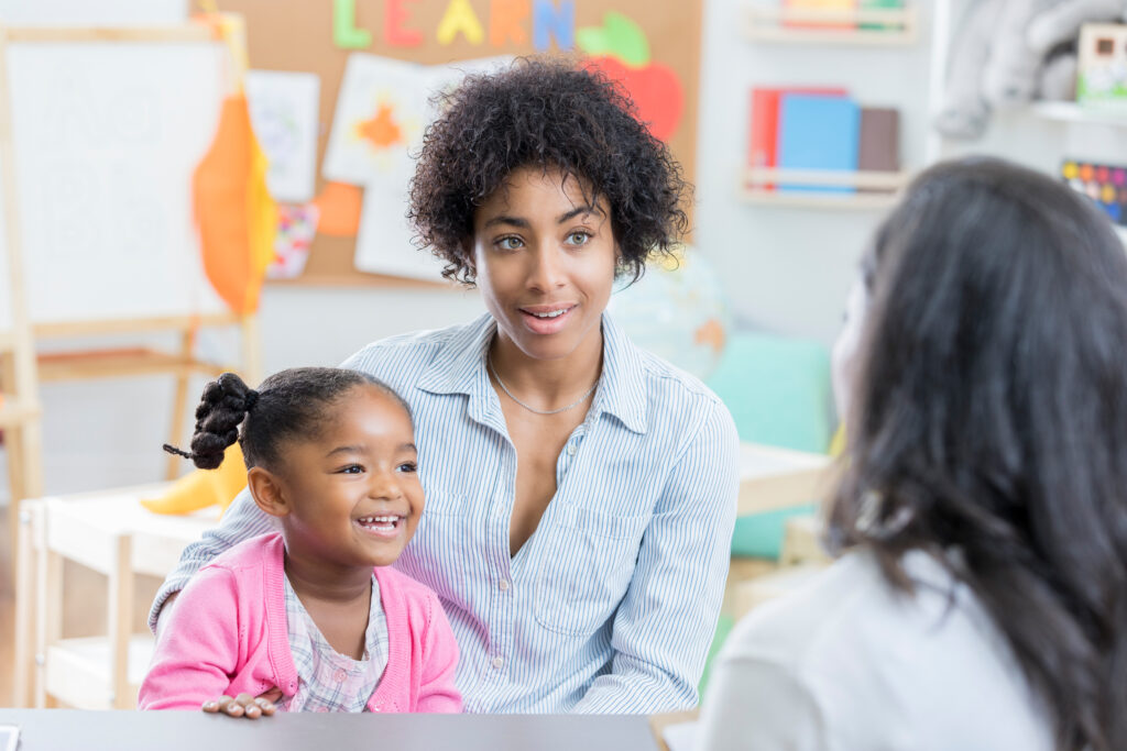 A young mother holds her preschool age daughter in her lap as she sits across a table from her daughter's new teacher for a meeting.