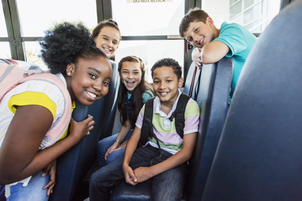 A group of multi-ethnic middle school students smile at the camera as they sit on a school bus.