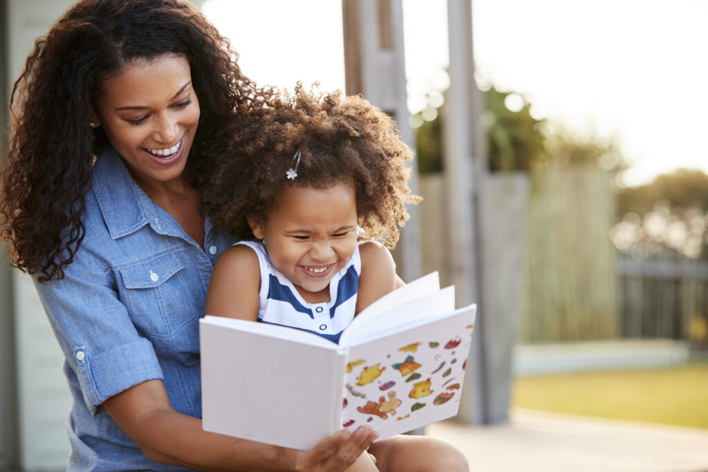 Young black girl reading a book while sitting on her mother's lap outside.