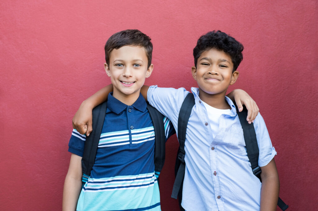 Portrait of multiethnic elementary school boys with backpacks, smiling with their arms around each other.