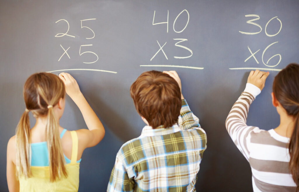 Three students stand facing a black board and are writing down their answers to multiplication math problems. 