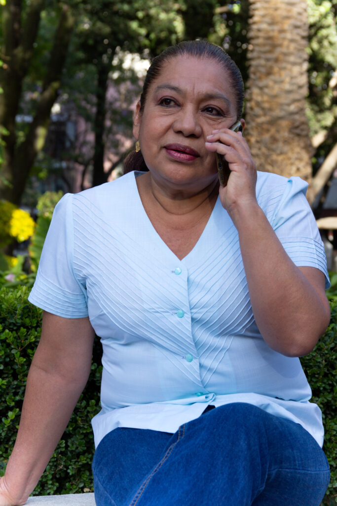 Mature Mexican woman talk on cell phone, sitting, in a park, with out of focus background of trees and green foliage. 