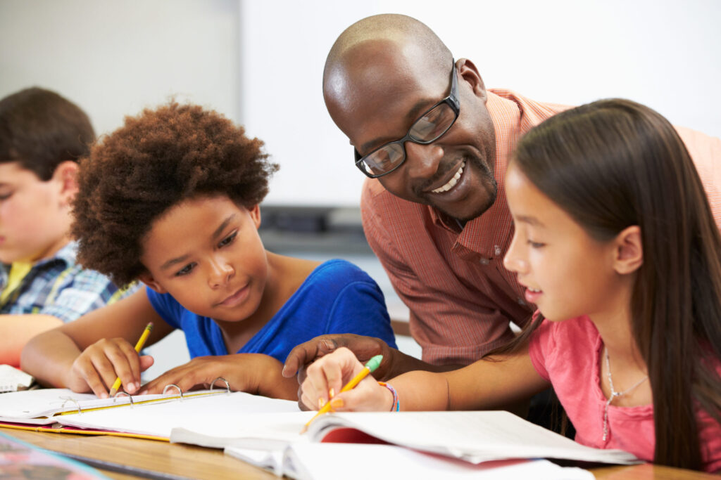 A black teacher smiles as he helps young multiethnic students with their work.
