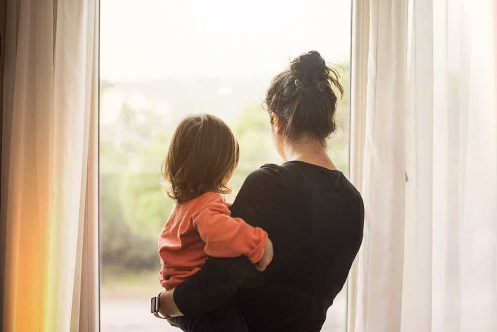 A mother holds her preschool child while looking out the window, her back facing the camera.