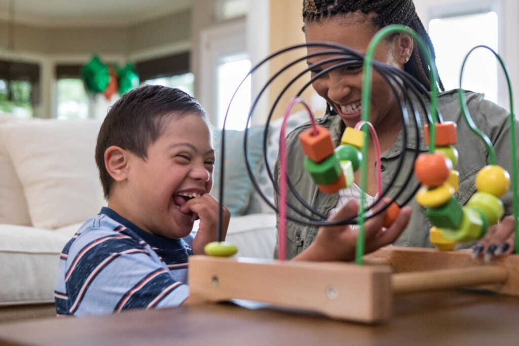 Smiling preschooler with down syndrome plays with a toy while his mother watches, also smiling.
