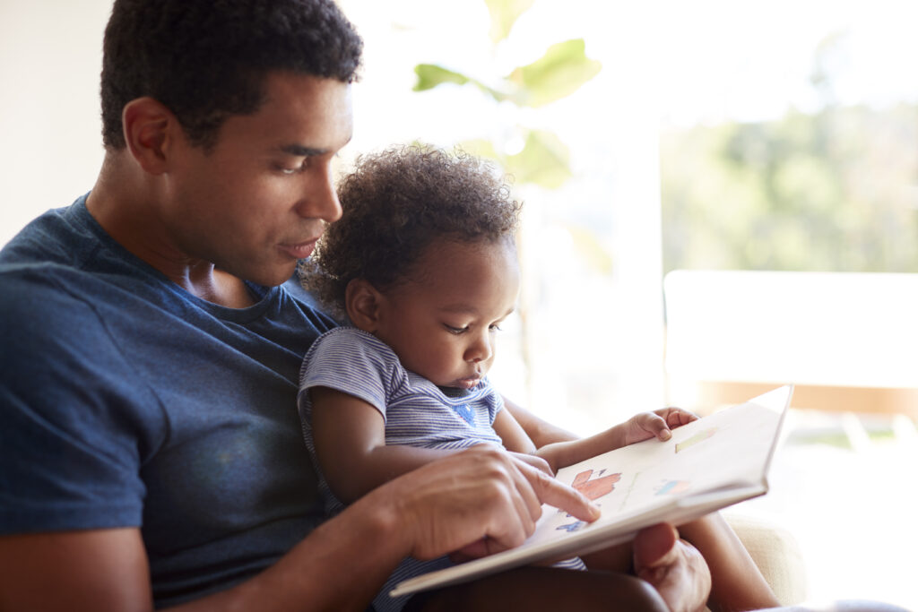 A black father reads a book with his two year old son at home.