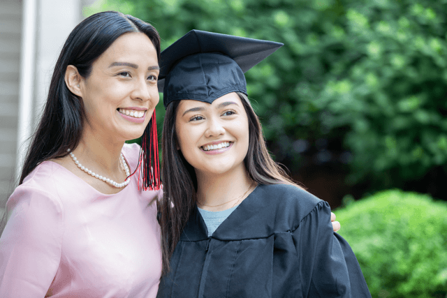 A mature Asian mother stands with her high school daughter at graduation. Both smile for a photo.