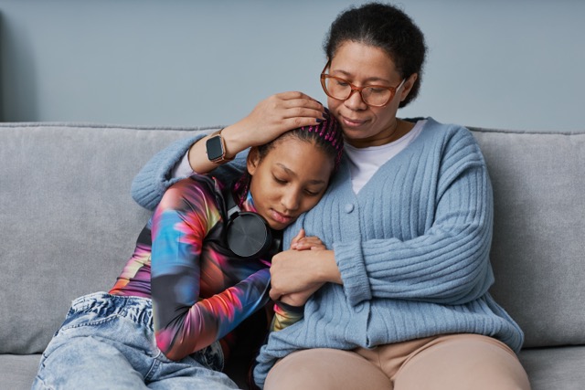 A mature black mother hugs her stressed teen daughter as they sit on the couch together. 