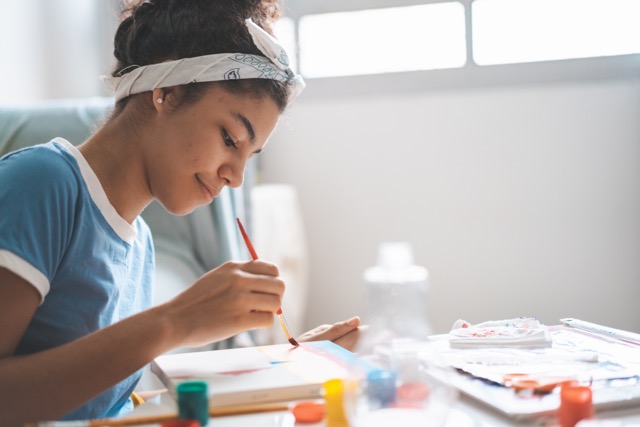 A teenage girl paints in her bedroom at home, appearing calm and content.