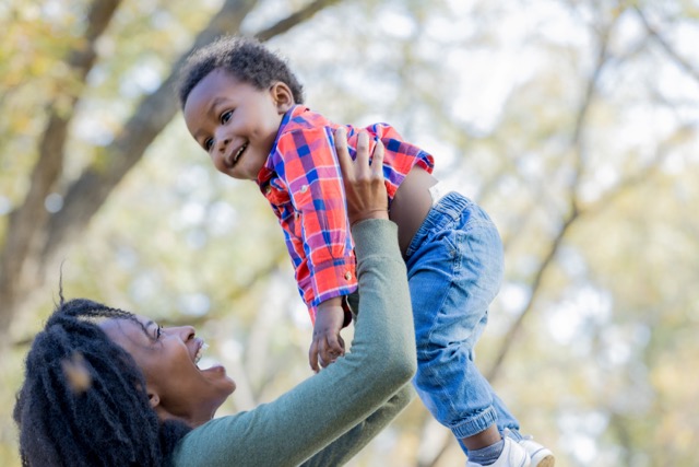 A smiling African American mother holds her baby boy high in the air on a sunny autumn day.