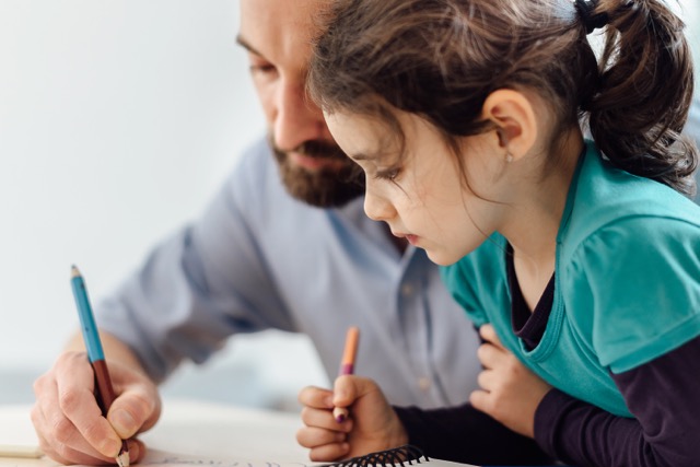 A Caucasian preschooler and her father draw together with colored pencils at a table.