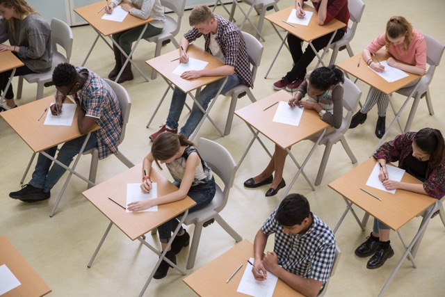 A classroom of high school students work on an exam. They are seated in individual desks in multiple rows. 