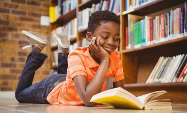 A Black boy lies on his stomach and reads a book at the library. 
