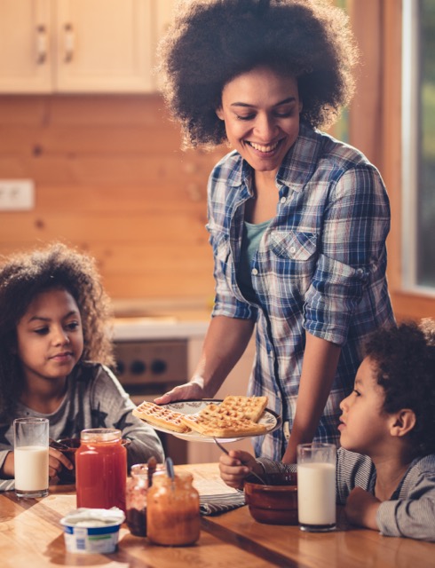 A smiling Black mother serves a meal to her two young children at home.